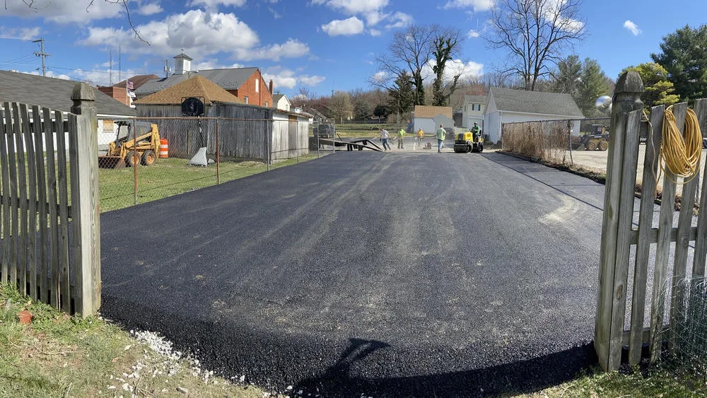 Five workers paving an asphalt lot on a sunny day. A yellow steamroller compacts the new surface.